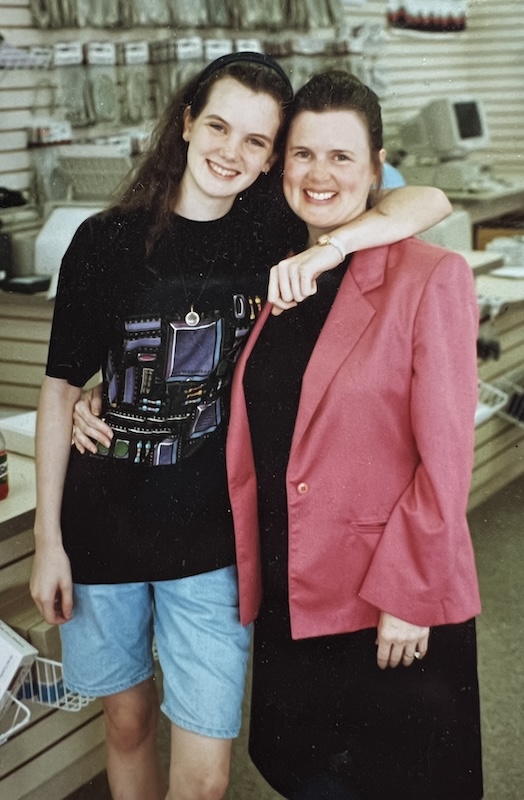 young woman with dark brown hair wearing jean shorts and a black t-shirt with circuit boards printed on it with her arms around the shoulders of an smiling older woman with dark brown hair in a bun, wearing a pink sport coat over a black top and skirt. they are clearly mother-daughter relation sharing a similar smile and clear blue eyes. they’re standing in a retail computer store circa 1995