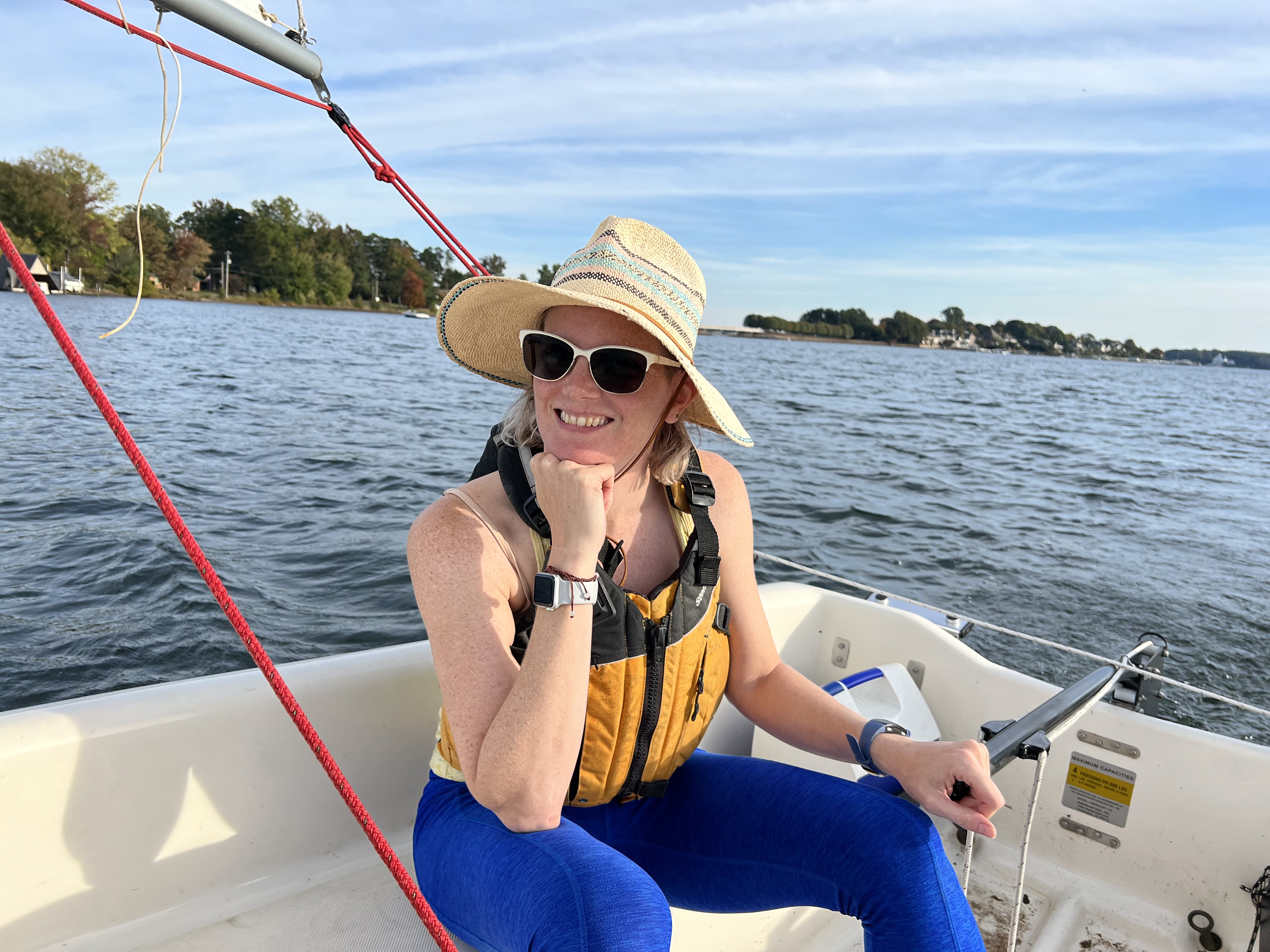 a woman sitting in the cockpit of a small sailing dinghy, which is on a lake, holding the tiller with her elbow on her knee and chin on her fist, smiling with a straw hat on and a life jacket. it’s a sunny day