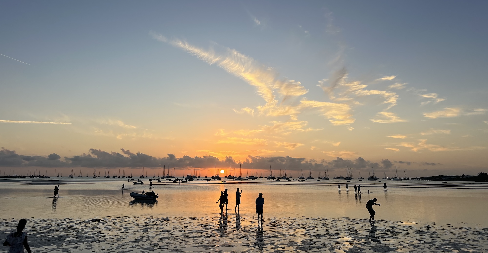 sunset over a beach at low tide with people playing in the foreground, dinghies and watercraft in the shallows, and numerous sailboats in the background