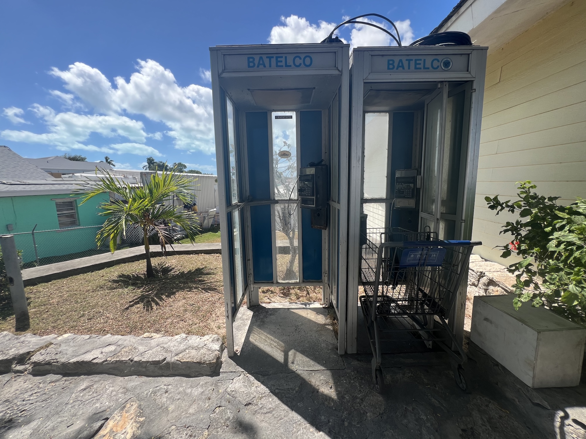 along a street, two old telephone booths on the right, one with a grocery cart parked inside it with a palm tree to the left and under a brilliant blue sky with white fluffy clouds