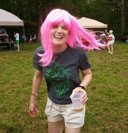 a young woman in her late 20s/early 30s, laughing while moving forward towards the camera. She's got long pink hair and is holding a beer, apparently at a festival